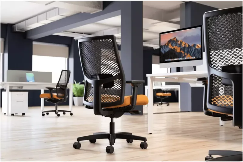 Black mesh office chairs with orange seats at white desks in an open workspace.